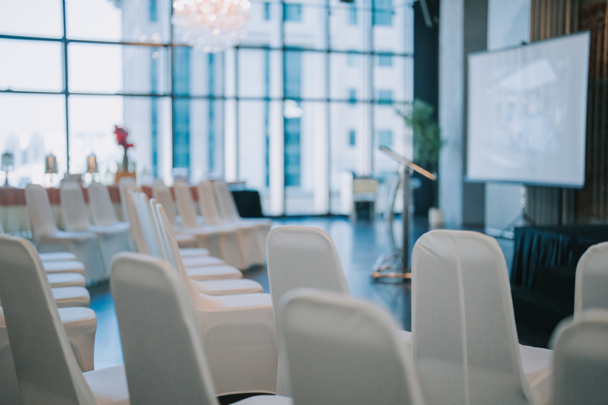 Empty chairs arranged in rows in a modern conference room with large windows, a whiteboard, and a podium.