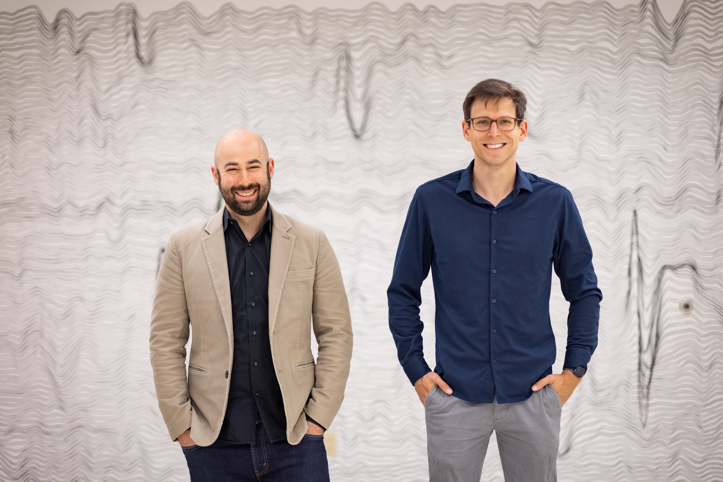 Dr. Brandman and Dr. Stavisky standing and smiling in front of a wall mural of thin wavy lines on a white background representing brain waves. Dr Brandman wears a tan blazer and Dr. Stavisky a navy button-down.