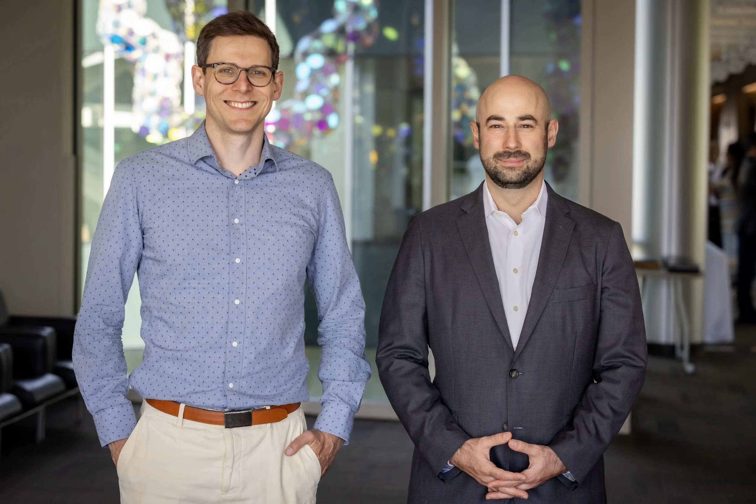 Dr. Sergey Stavisky and Dr. David Brandman stand in a modern hospital lobby with glass walls with colorful reflection in the background, smiling at the camera.