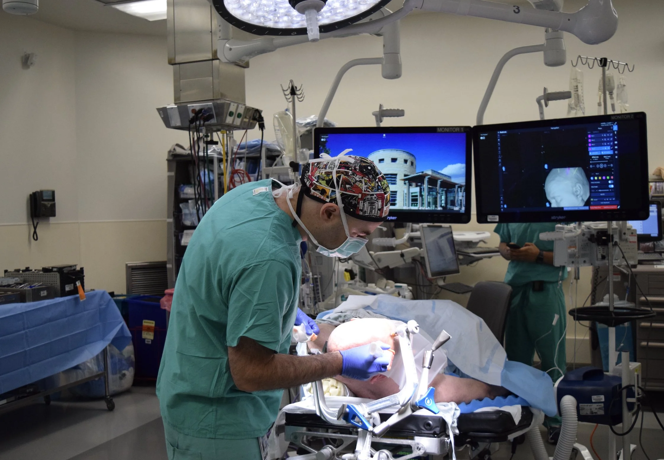 Dr. David Brandman is performing neurosurgery in an operating room, with two monitors displaying medical images and various surgical equipment around them.