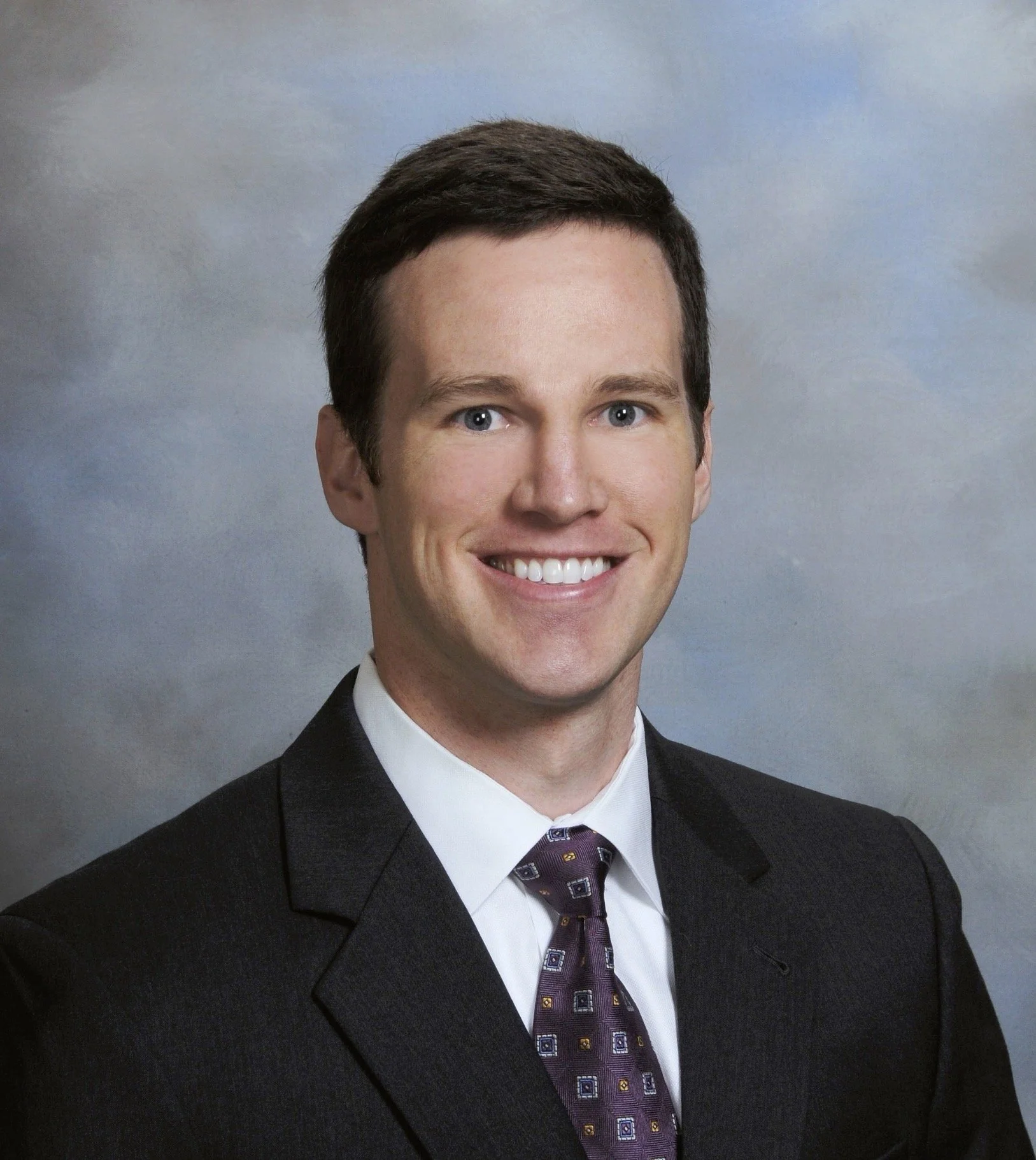 Professional headshot of a man in a dark suit, white shirt, and patterned tie, smiling against a cloudy sky background.