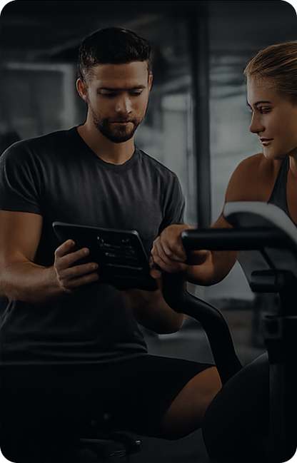 A man and woman at a gym, looking at a smartphone together near a treadmill.