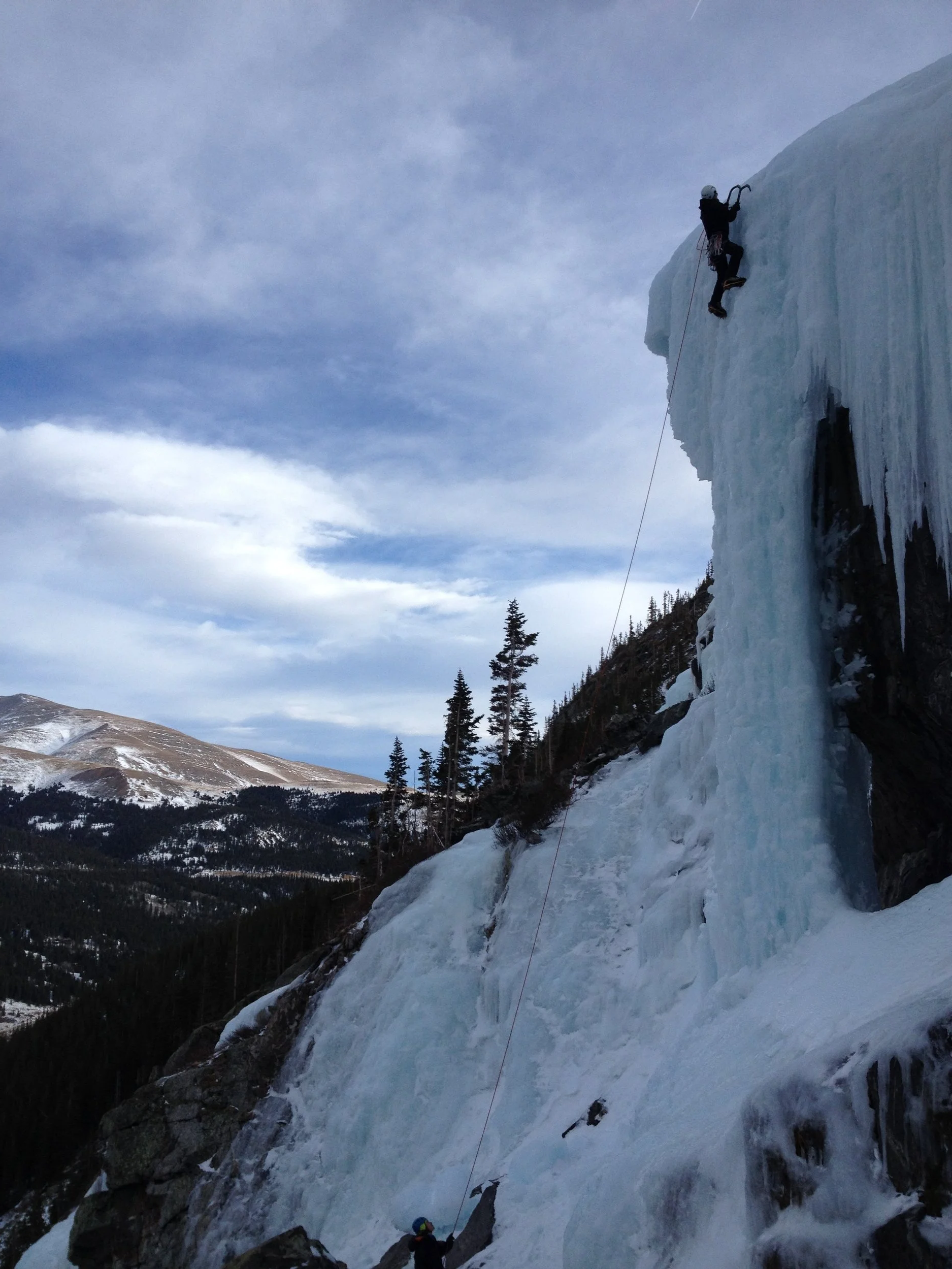 Dr. Jackson Bell ascending a frozen waterfall with a safety rope, while another climber below assists.