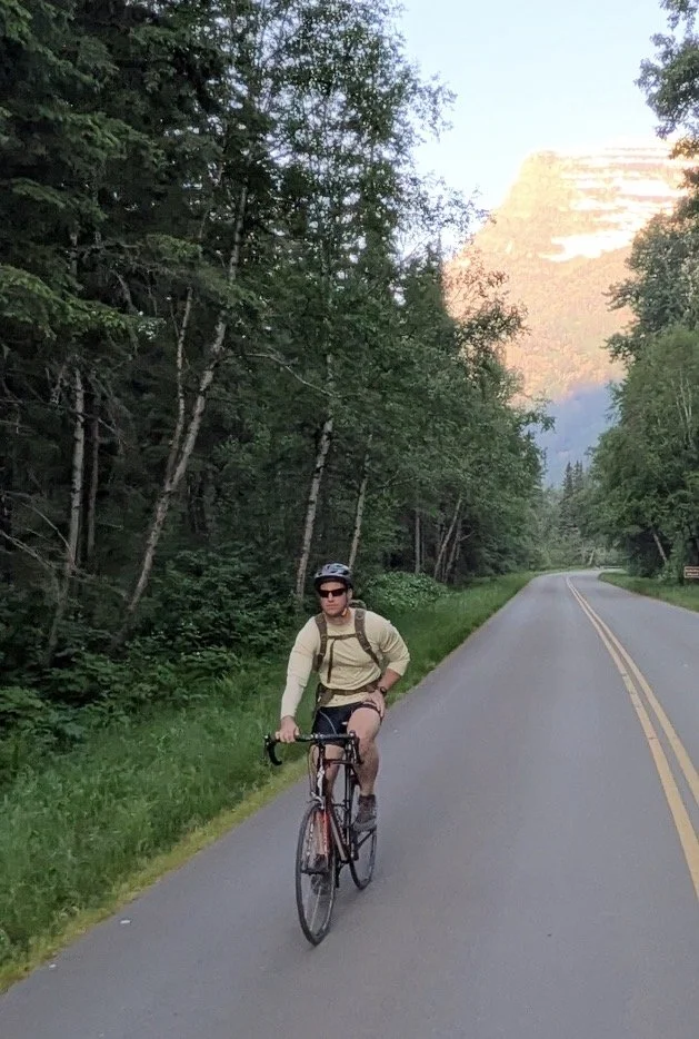 A man cycling on a paved road surrounded by trees, with a mountain in the background illuminated by the sunlight.