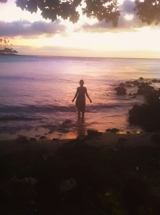 Person standing in shallow ocean water at sunset, with clouds and trees in the background.