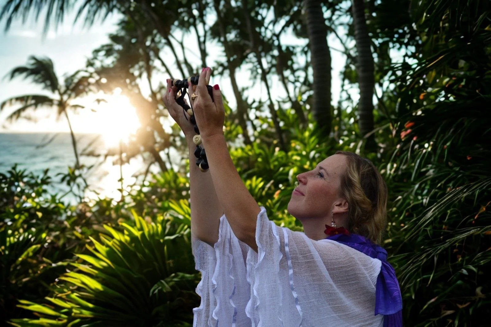 Woman chanting and holding a black and white beaded necklace outdoors at sunset, surrounded by green tropical plants and palm trees.