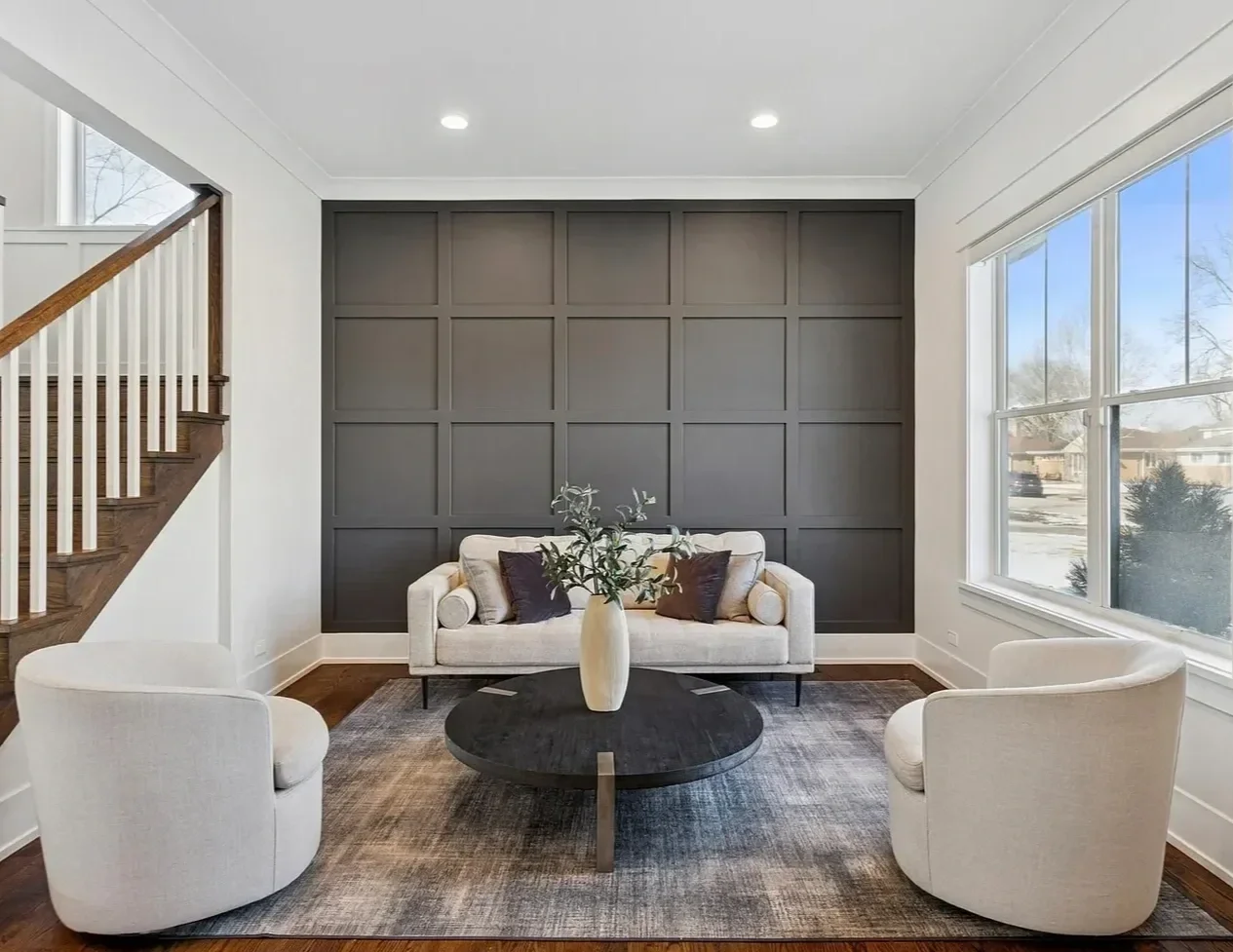 Living room with modern white sofa, two white armchairs, dark wood coffee table with a vase of greenery, dark paneled accent wall, large windows, and hardwood floors.