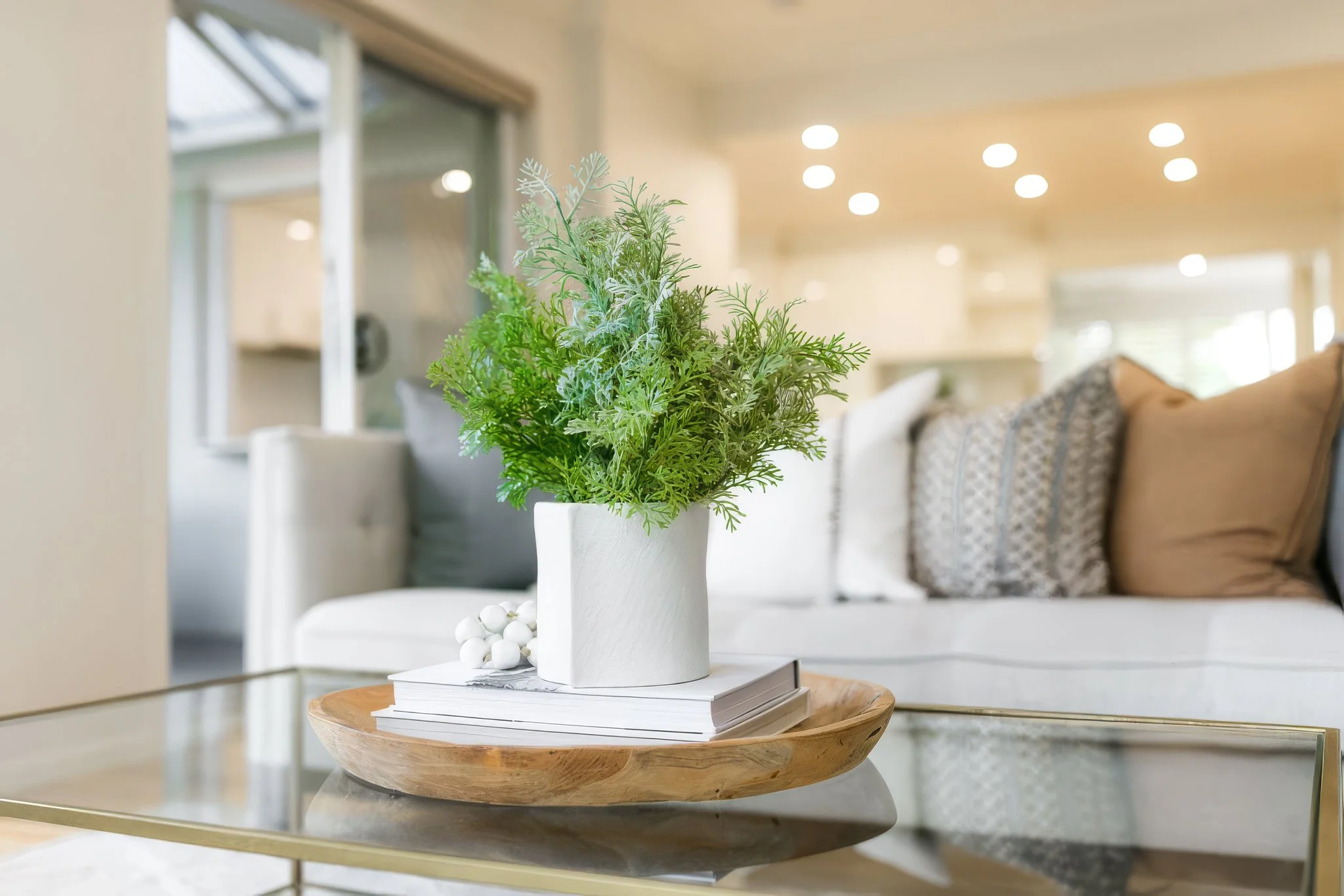 A white ceramic vase with green foliage sitting on a stack of white books on a wooden tray in a bright living room with neutral tones.