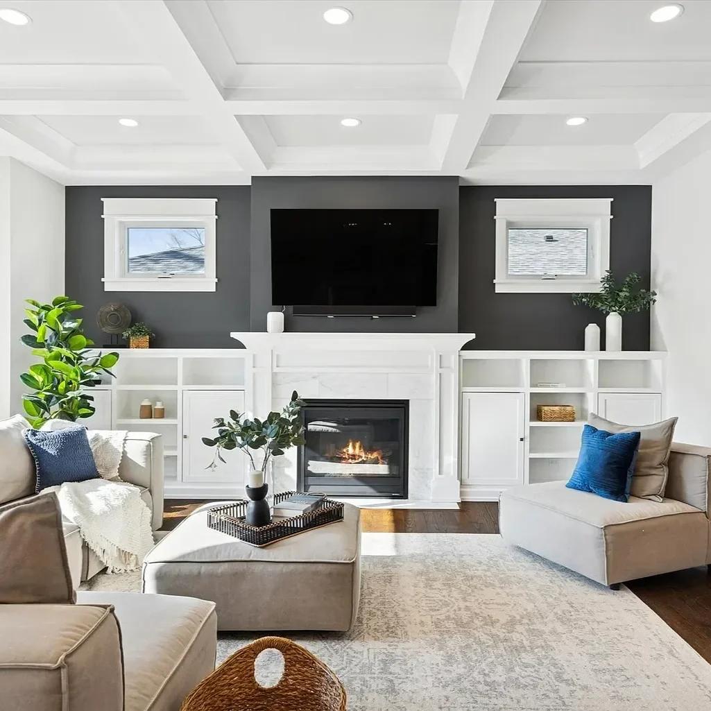 Living room with white walls and ceiling, dark gray accent wall with a mounted TV, fireplace, two small windows, white built-in shelves, beige sofas with blue cushions, potted plants, and a beige area rug.