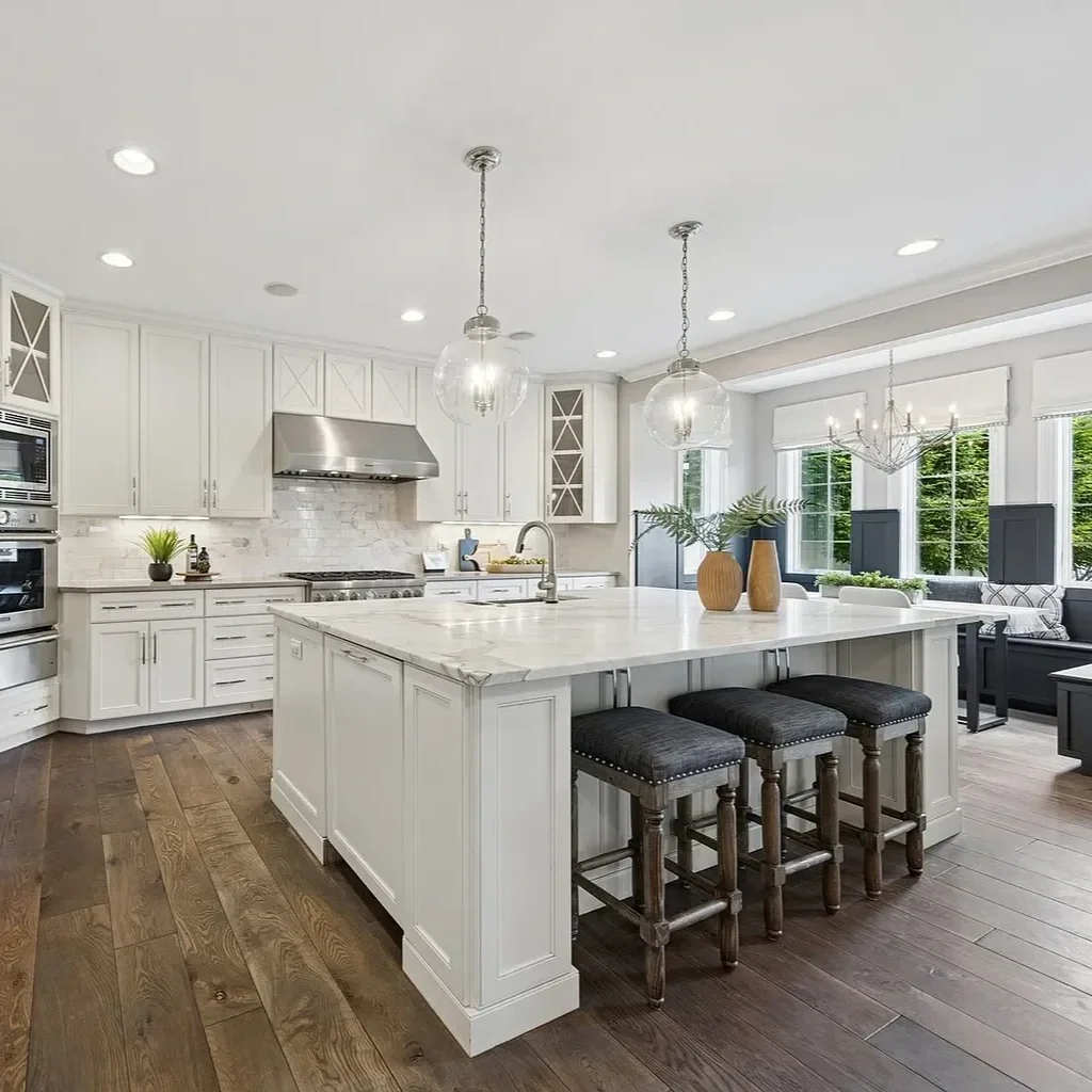 Bright modern kitchen with white cabinets, marble island, wooden flooring, stainless steel appliances, glass pendant lights, and large windows with greenery outside.