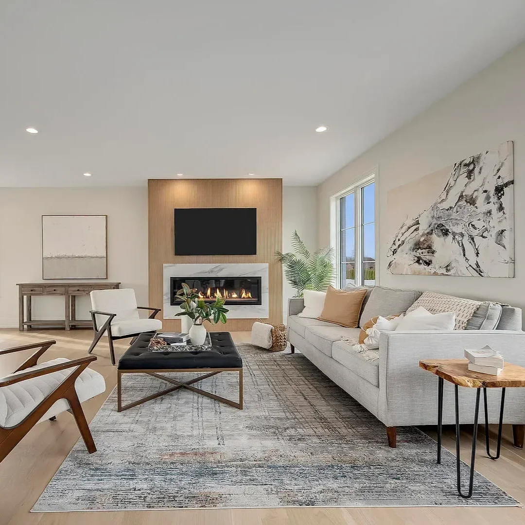 Modern living room with white sofa, armchairs, a black tufted coffee table, a gray rug, fireplace, wall-mounted TV, large window, artwork, and indoor plants.