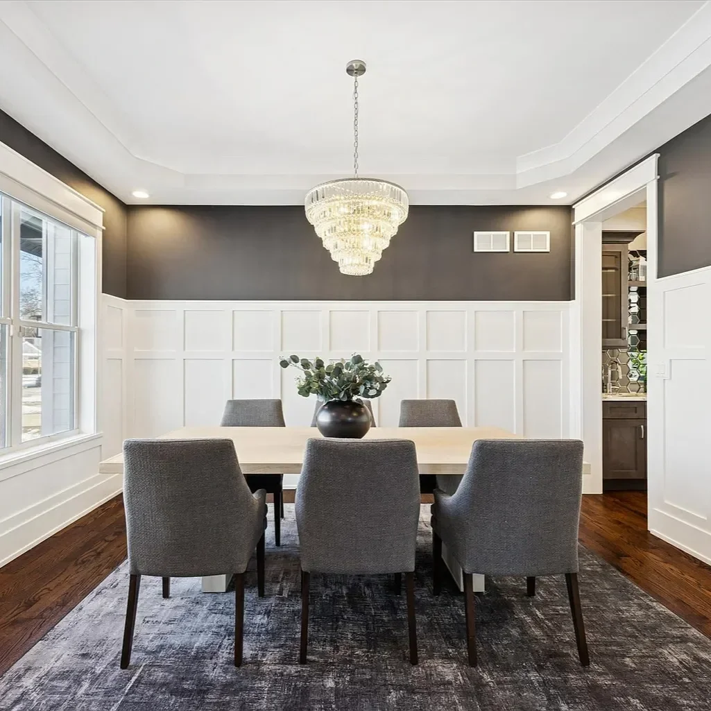 A dining room with a modern chandelier, six gray upholstered chairs around a light wooden table, a dark gray accent wall with white paneling, a large window, and hardwood floors.