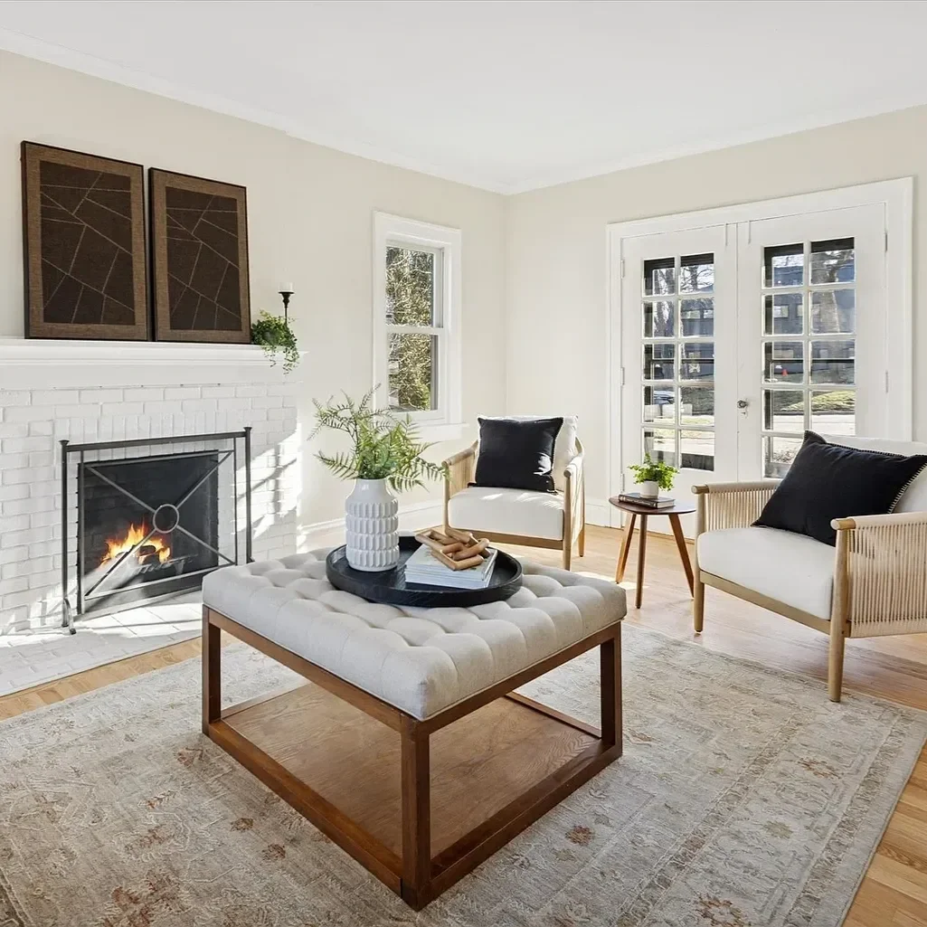 Bright living room with white walls, a white brick fireplace, and a fire burning. There are two armchairs with black pillows, a tufted beige ottoman with a tray holding a plant, books, and decorative items. French doors and a window let in natural li