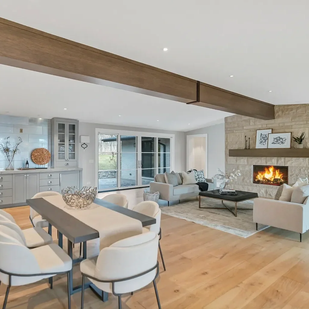 Living room with a dining area, light-colored sofas, a fireplace, a wooden beam on the ceiling, large sliding glass doors, and neutral decor.