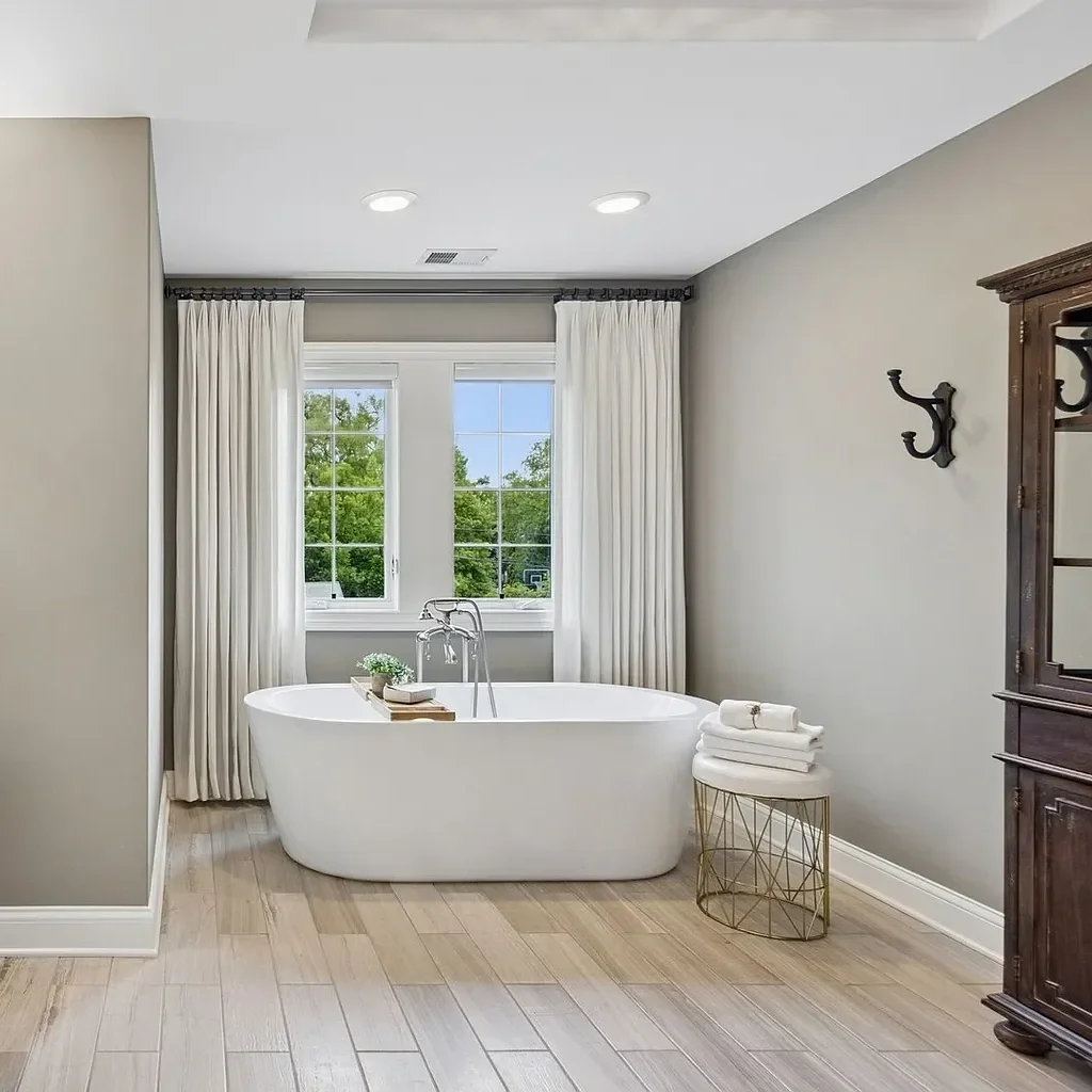 A modern bathroom with a white freestanding bathtub near two large windows, white curtains, a small side table with rolled towels, a dark wooden cabinet, and a decorative wall hook.