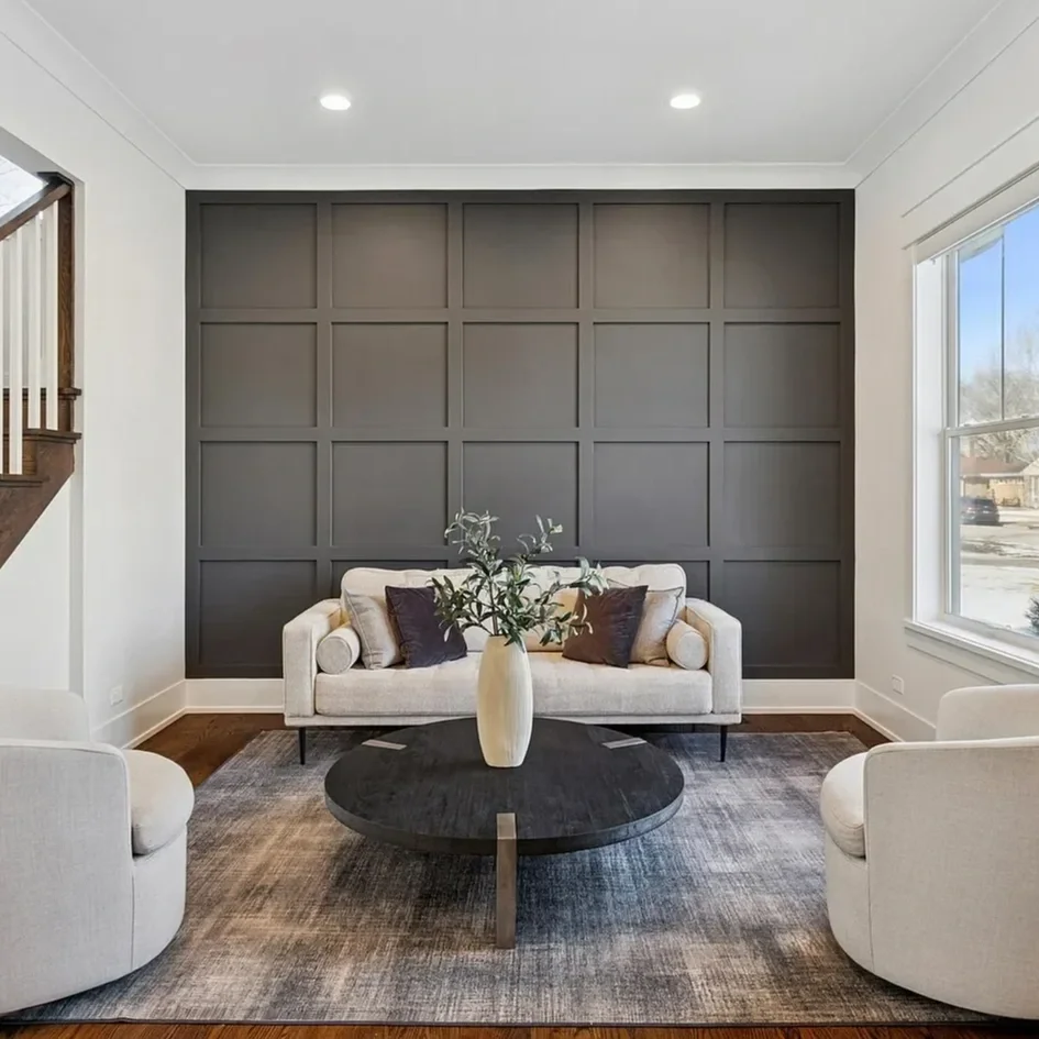 Living room with a gray paneled accent wall, a white sofa with dark and light pillows, a round black coffee table with a white vase and green foliage, two white armchairs, a window to the right, and a staircase to the left.
