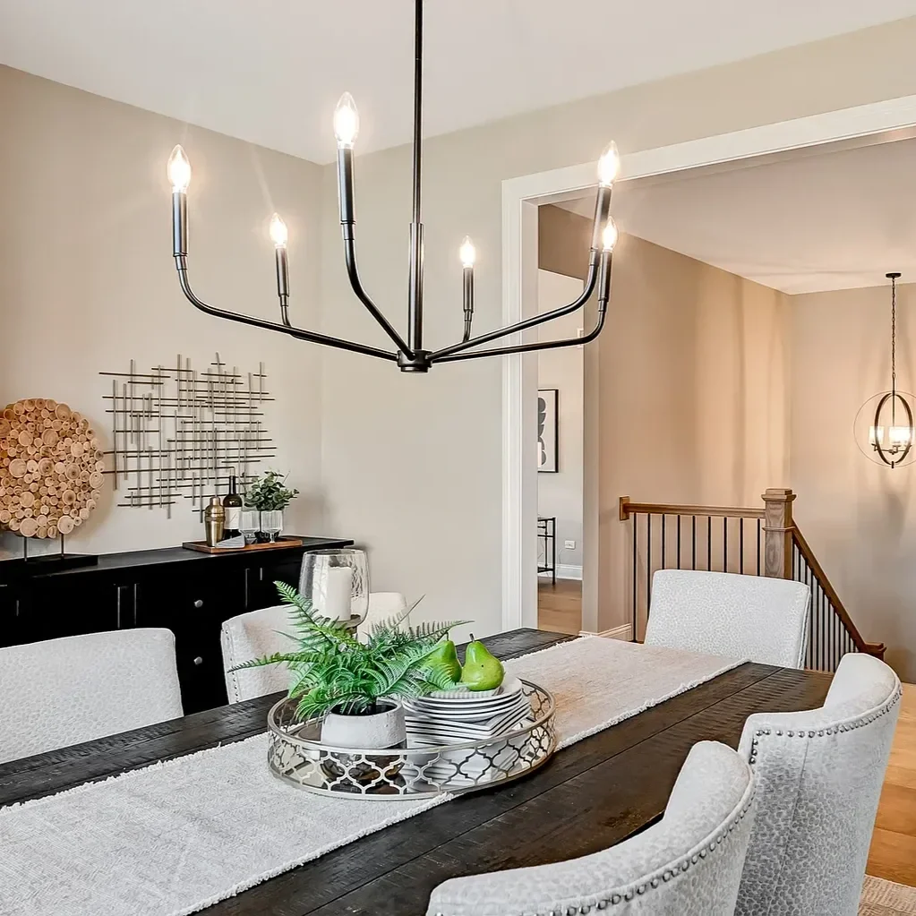 A dining room with a dark wood table, white upholstered chairs, a modern black chandelier with candlestick bulbs, a black sideboard with decorative art and plants, and a staircase with wooden railing in the background.