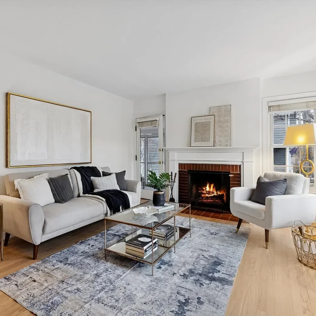 A cozy living room with a white sofa, an armchair, a glass coffee table, and a fireplace with a fire burning. There are art pieces on the wall, a patterned rug, a plant, and a side table with a lamp. Large windows let in natural light.