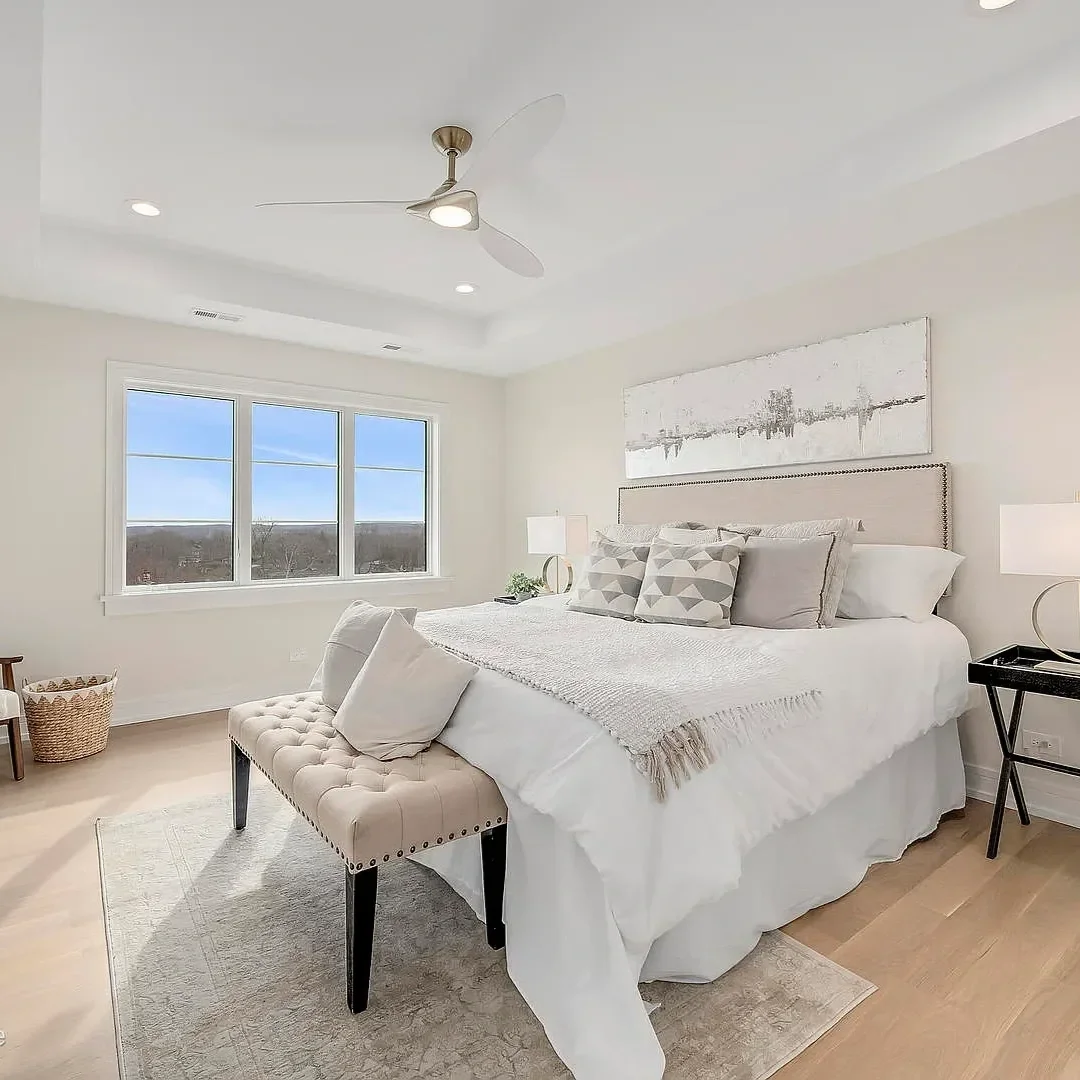 A bright bedroom with a king-size bed, white bedding, decorative pillows, nightstands with lamps, a window showing a clear sky, and a ceiling fan with light.