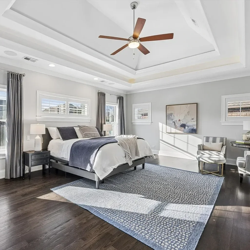 A modern bedroom with a large bed, nightstands with lamps, two windows with gray curtains, a patterned area rug, a ceiling fan, and a chair near the wall. The room has light-colored walls and dark hardwood floors.
