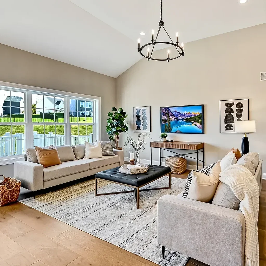 Living room with beige sofas, a black leather ottoman, a large window, a wall-mounted TV, and modern artwork, with a chandelier hanging from the ceiling.