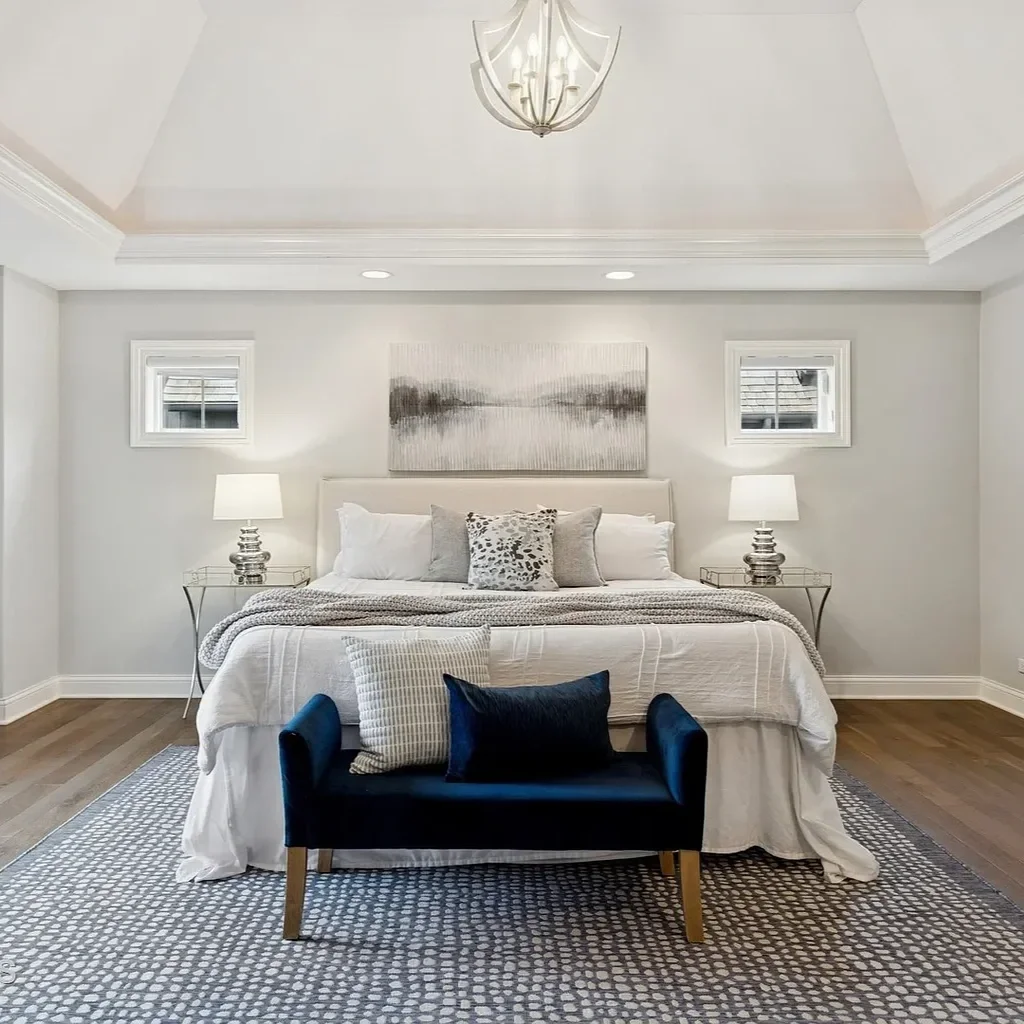Modern bedroom with white walls, a bed with white bedding and multiple pillows, two side tables with lamps, a black bench with pillows at the foot of the bed, and two small windows behind the bed, with a chandelier hanging from the ceiling.