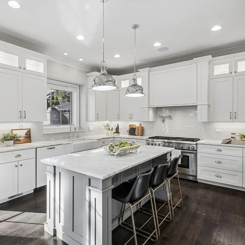 Modern white kitchen with marble island, black chairs, and stainless steel appliances, brightened by pendant and recessed lighting.