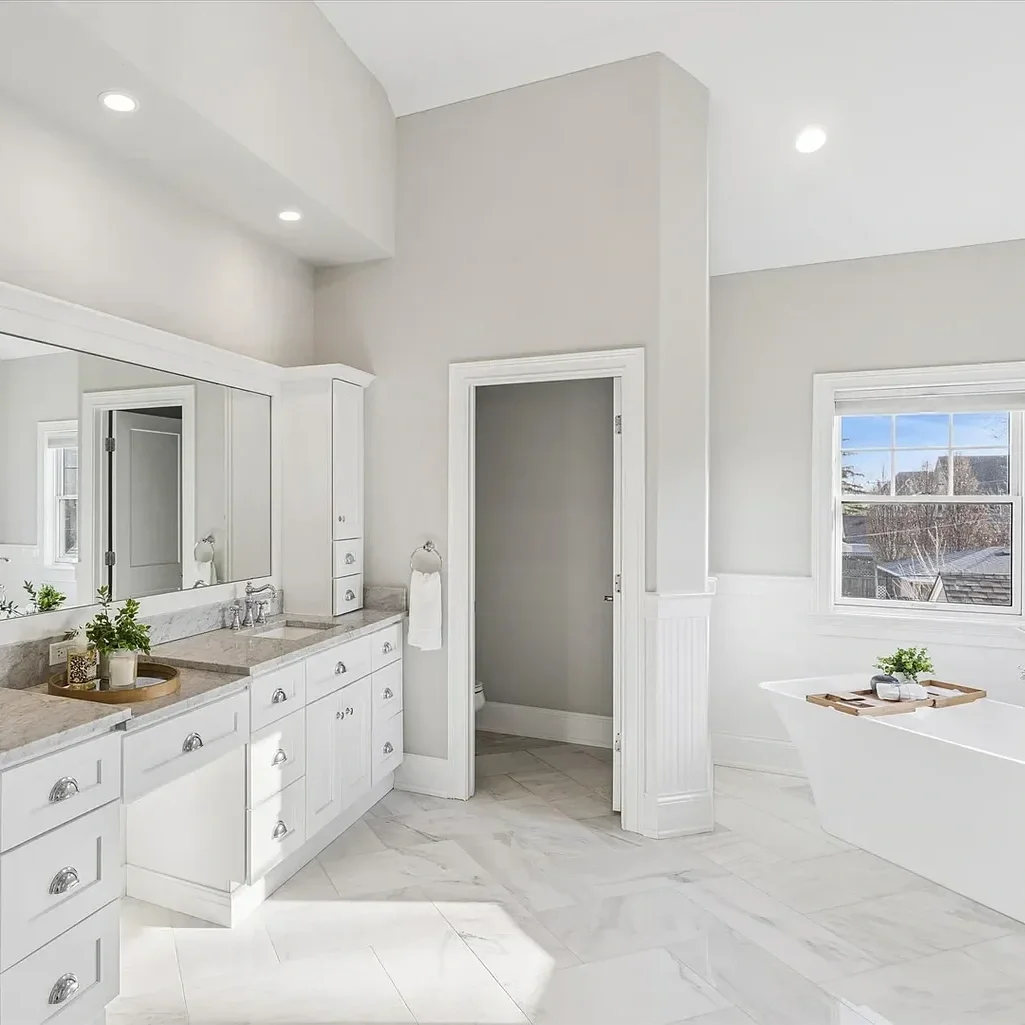 Bright bathroom with white cabinets, marble countertops, and a bathtub near a window.
