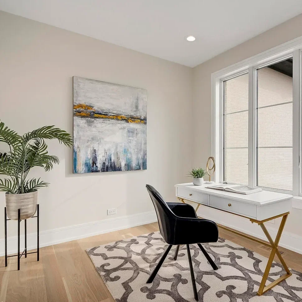 A home office with a white desk, a black chair, a window, a potted plant, and an abstract painting on the wall.