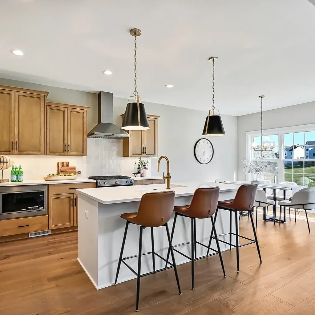 Modern kitchen with wooden cabinets, white countertop island with three brown barstools, black pendant lights, stainless steel range hood, microwave, and a large window in the background letting in natural light.