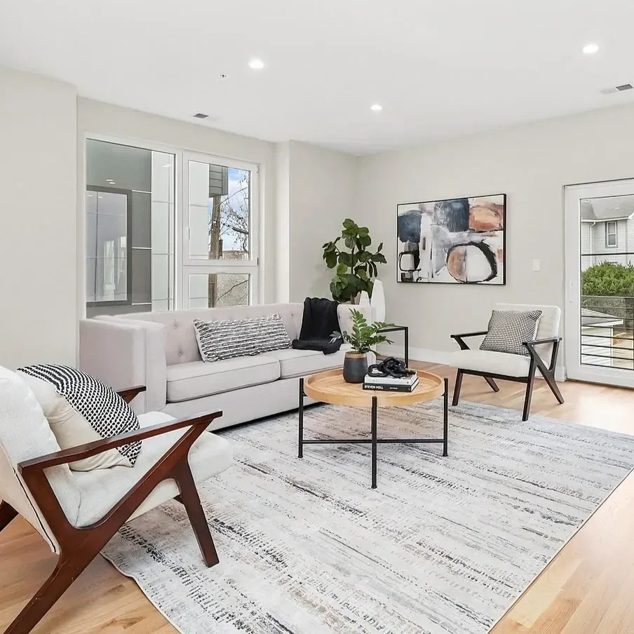 Modern living room with white sofa, black and white patterned pillows, abstract wall art, large window, sliding door, potted plant, round coffee table with books and decor, armchair with wooden frame, rug, and hardwood floors.