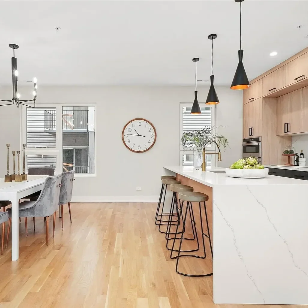 Modern kitchen and dining area with light wood flooring, white walls, black pendant lights, a marble island with stools, a wooden dining table with four grey chairs, and a wall clock.