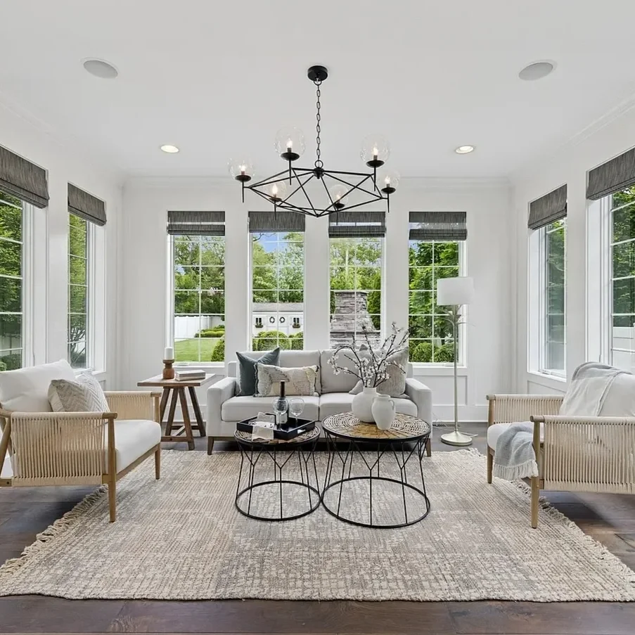 Bright living room with white walls, multiple large windows with gray blinds, a white sofa, two white armchairs with wooden arms, and two black wireframe coffee tables with white vases and decorative branches, a floor lamp, a chandelier, and a beige 