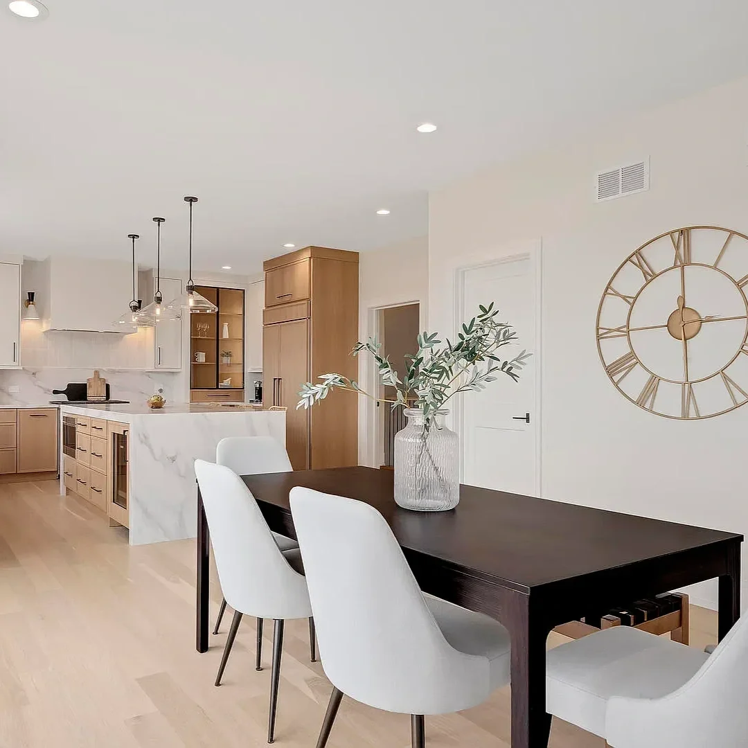 Modern open-concept kitchen and dining area with a dark wooden table, white chairs, a large wall clock, and a vase with greenery on the table.