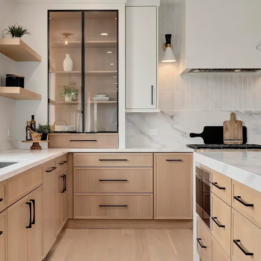 Modern kitchen with light wood cabinets, white quartz countertops, a black glass-front cabinet, open shelves with plants and decorative items, a marble backsplash, black hardware, a wall-mounted light fixture, and minimal kitchen accessories.