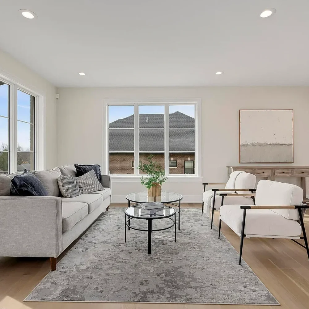 Bright living room with a beige sofa, two white armchairs, a gray rug, a round glass coffee table, and a window showing a brick building outside.