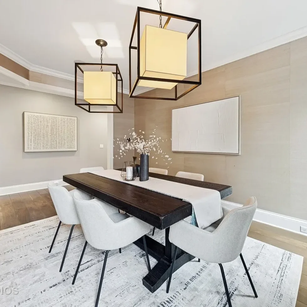 A modern dining room with a dark wooden table, six white upholstered chairs, and two geometric pendant lights hanging from the ceiling. There are two large abstract art pieces on the walls, a vase with flowers on the table, and a white area rug on a 