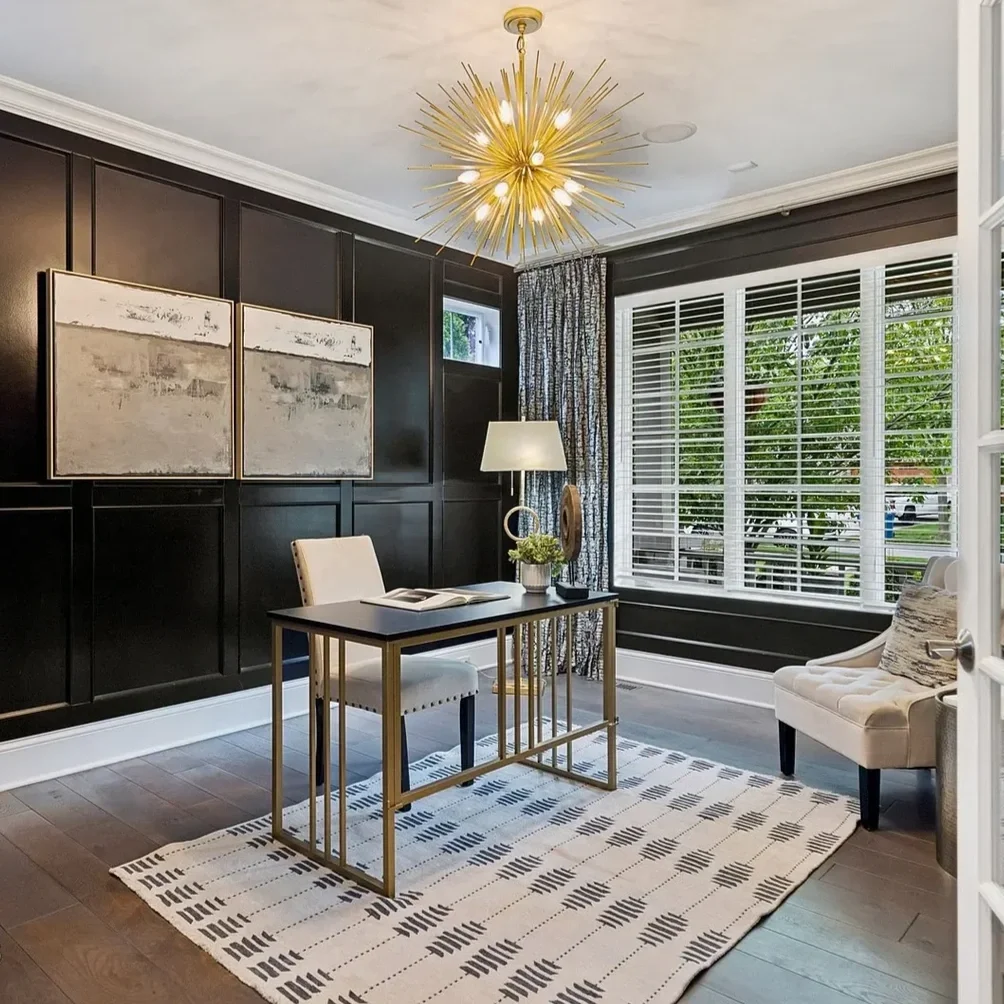 Modern home office with dark accent wall, large window with white blinds, white desk with gold frame, white chair, beige armchair, abstract artwork, gold starburst chandelier, patterned rug, and decor items.