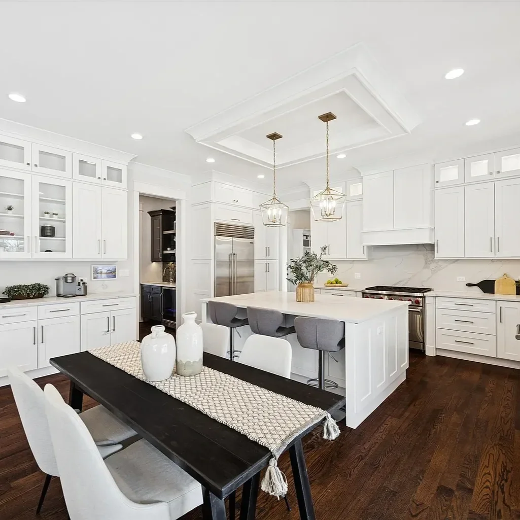 Modern white kitchen with dark hardwood floors, white cabinetry, a central island with seating, stainless steel appliances, and a dark wood dining table with white chairs.
