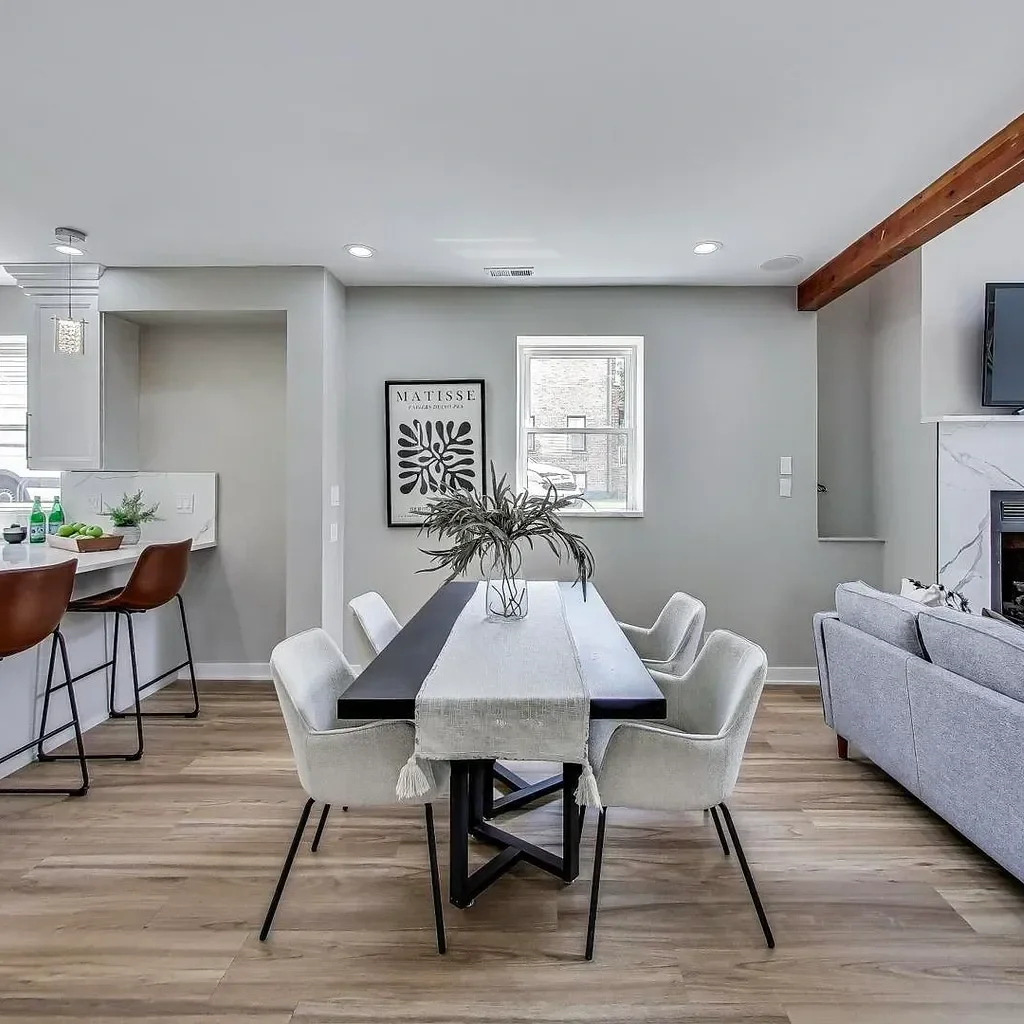 Modern dining area with light gray walls, a black dining table with a beige table runner, six beige upholstered chairs, a large floral arrangement in the center, a framed black and white art piece, a window, and a view of a gray sofa and fireplace on