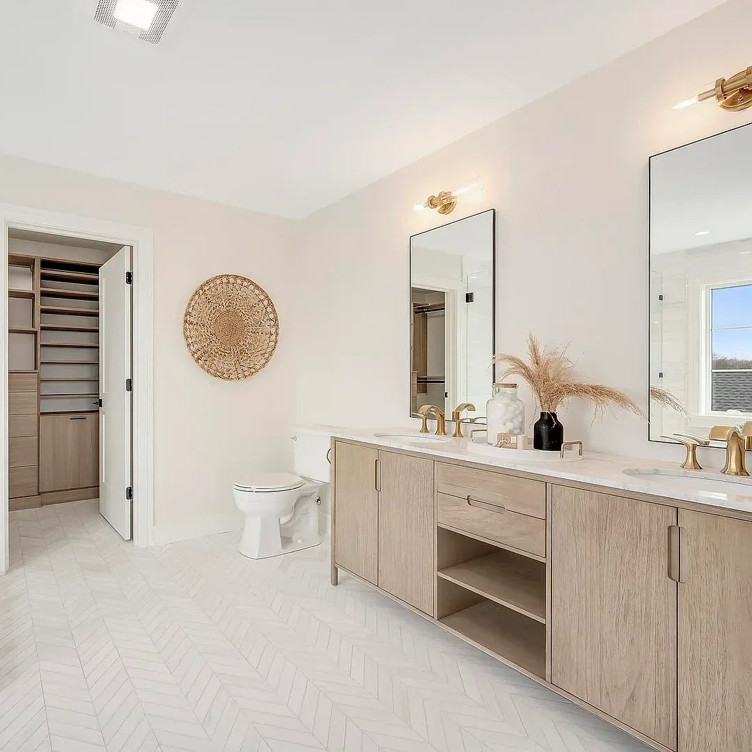 Modern bathroom with dual sinks, large mirrors, gold fixtures, and a light wood vanity. There is a toilet and a walk-in closet with built-in shelving visible through an open door. Decor includes a black vase with dried pampas grass and a decorative w