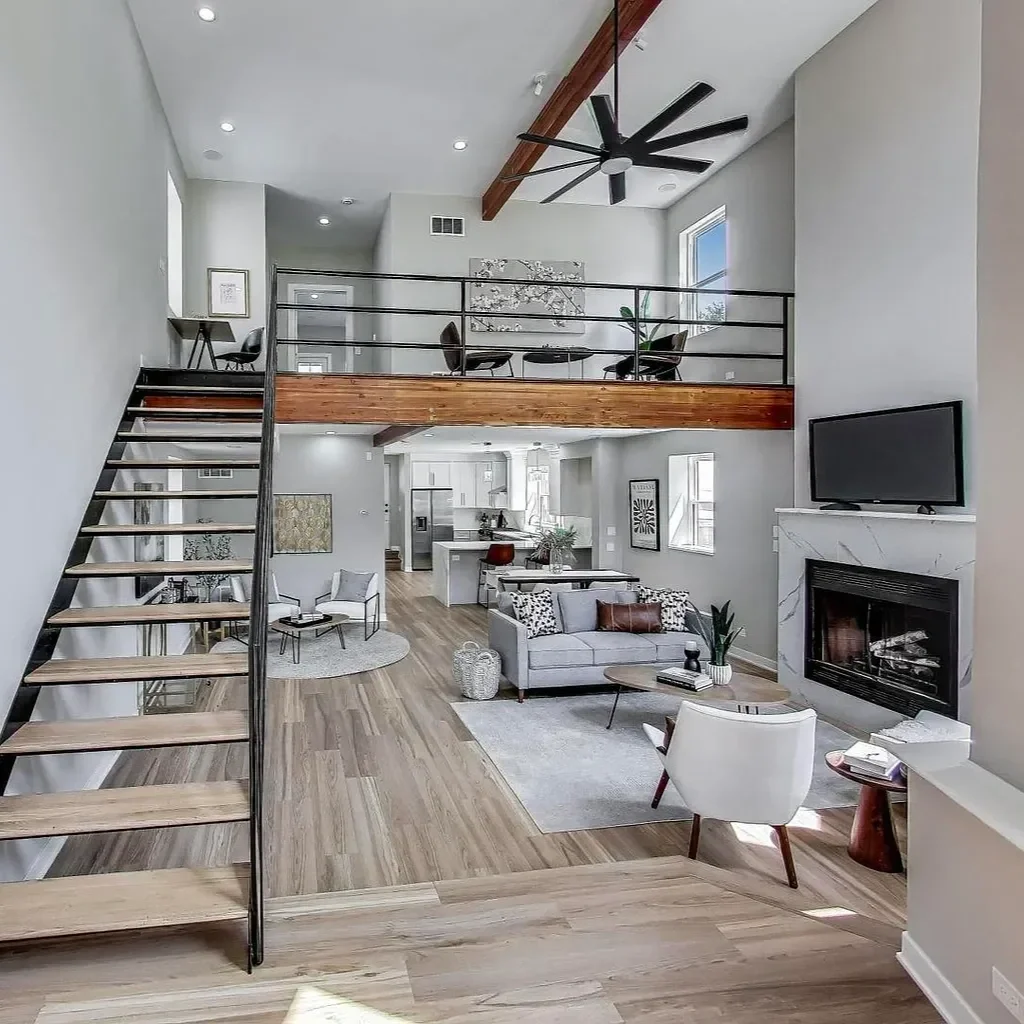 Bright modern living room with hardwood floors, a fireplace, a gray sofa, white armchair, coffee table, wall-mounted TV, and large windows. An upstairs balcony with a black railing overlooks the space.