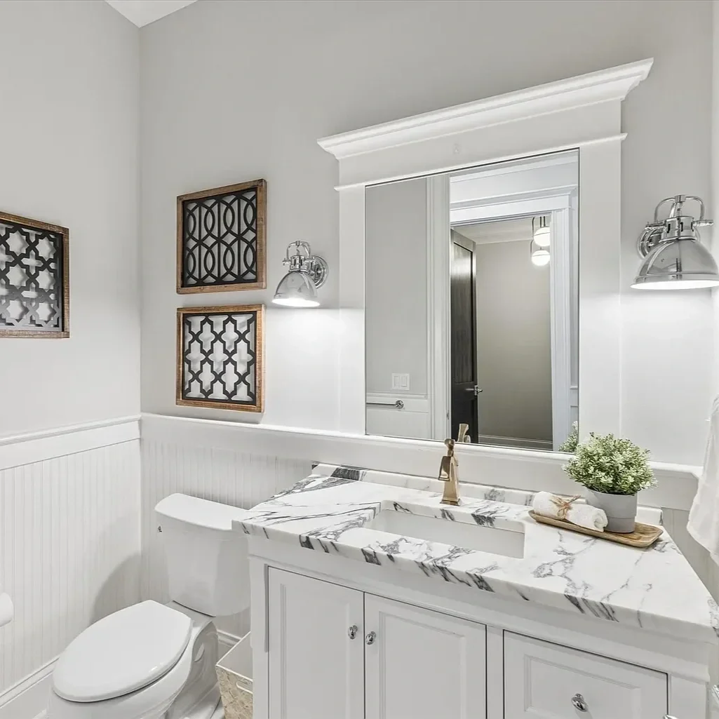 A modern bathroom with a white marble countertop and sink, a white toilet, a large mirror surrounded by decorative trim, two wall-mounted chrome lamps, and framed black lattice wall art.