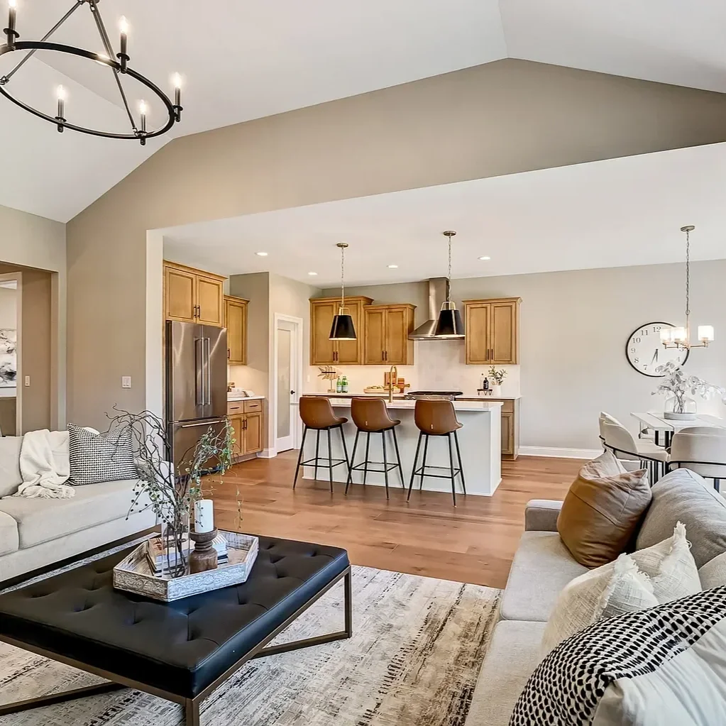 Open-concept living room and kitchen with beige walls, hardwood floors, and modern furnishings. Living room has a beige sofa with pillows, a black coffee table with decorative branches, and a chandelier. Kitchen features wooden cabinets, stainless st