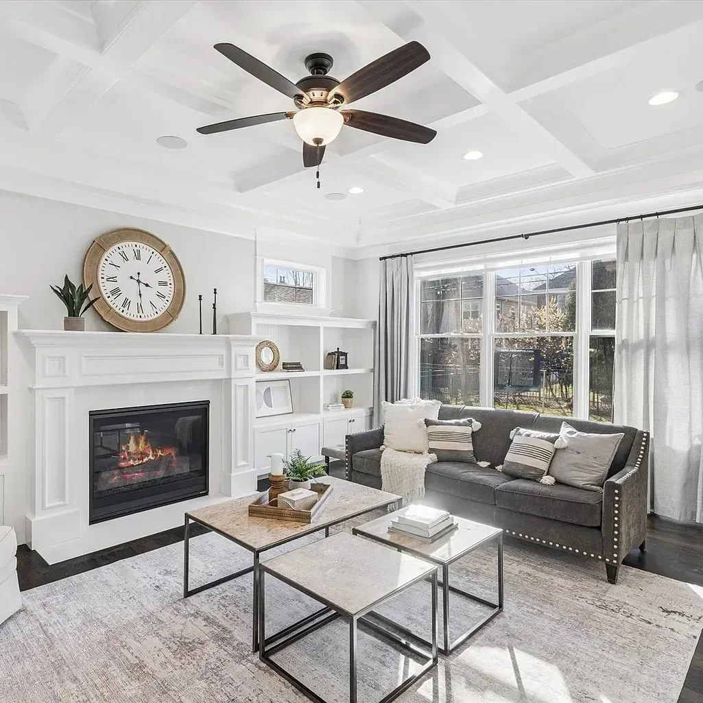 Bright living room with white walls, ceiling with recessed lighting, a ceiling fan, large window with curtains, a gray sofa with pillows, a coffee table with books, and a fireplace with a large round clock above it.