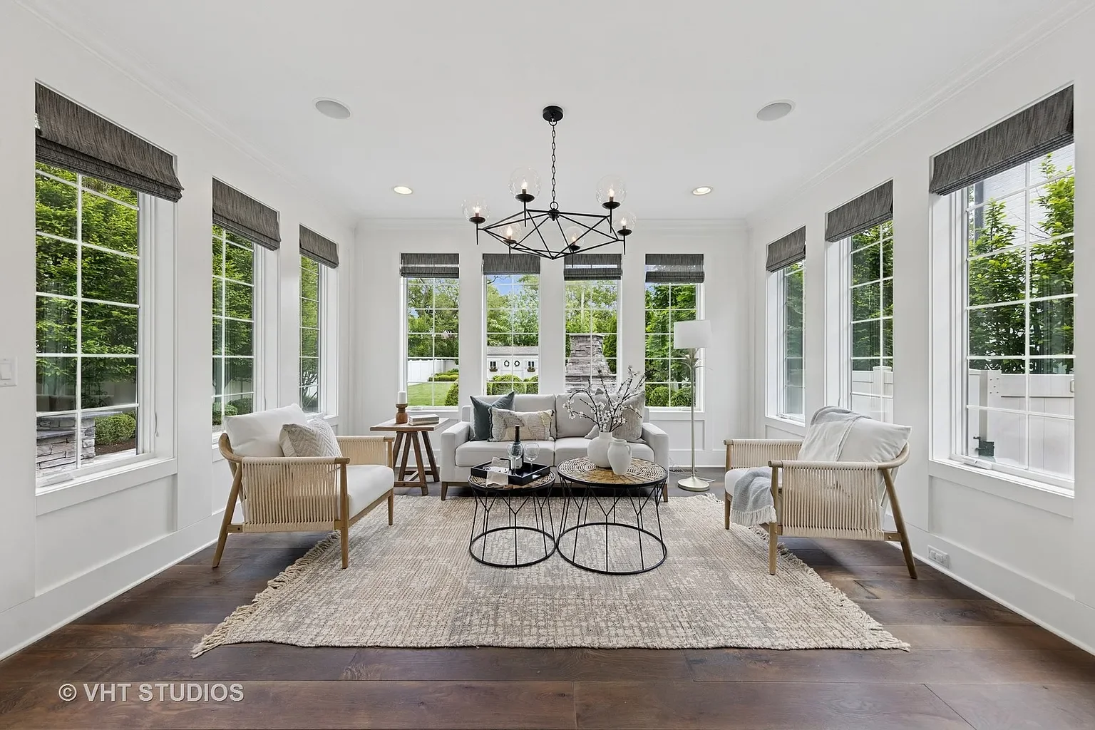 Bright living room with large windows, white walls, a white sofa, two rattan armchairs, a beige rug, and black metal coffee tables with decorative vases and glasses.