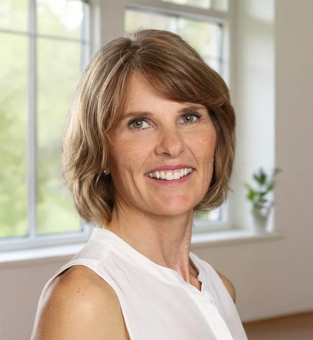 A woman with shoulder-length light brown hair, wearing a white sleeveless blouse, smiling, standing indoors near a window with green trees outside and a small potted plant on the windowsill.