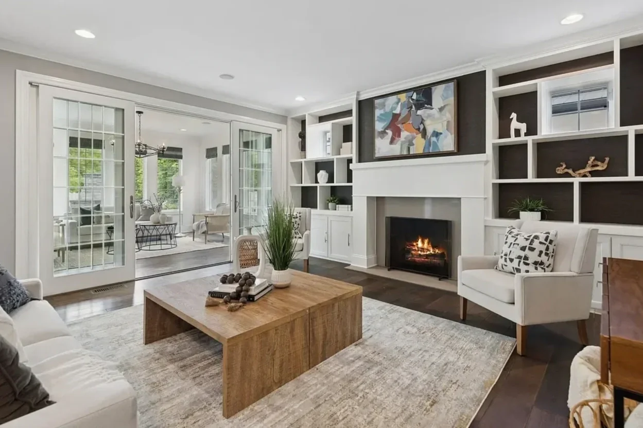 Living room with white fireplace, built-in shelves, and sliding glass doors leading to a sunroom, decorated with modern art, plants, and furniture.