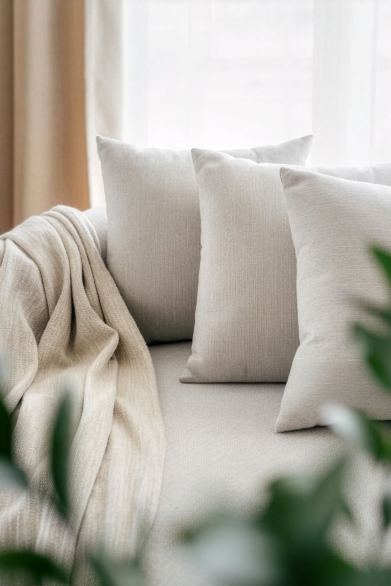 Close-up of beige pillows and a beige blanket on a light-colored sofa near a window with sheer curtains.