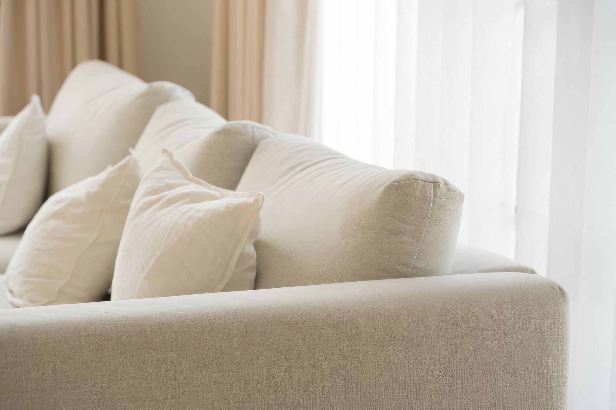 A close-up view of a cream-colored sofa with multiple white pillows, set in front of sheer curtains with natural light filtering through.